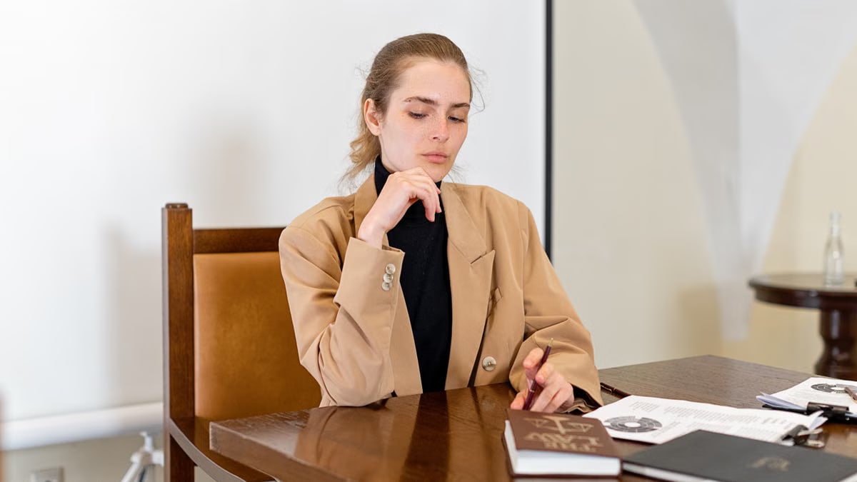 Caucasian woman in a brown blazer sitting at a desk with documents, looking thoughtful.